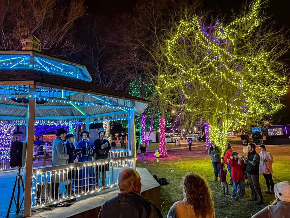 A pavilion lit up at with blue string lights with carolers on the inside next to lit up tree
