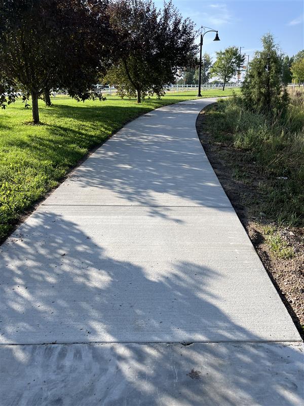 Concrete walking path curving through a grassy park with trees and a lamppost.