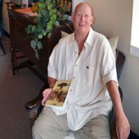 Sean Scott, author of Around Frederick sitting in a chair wearing a white shirt holding his book
