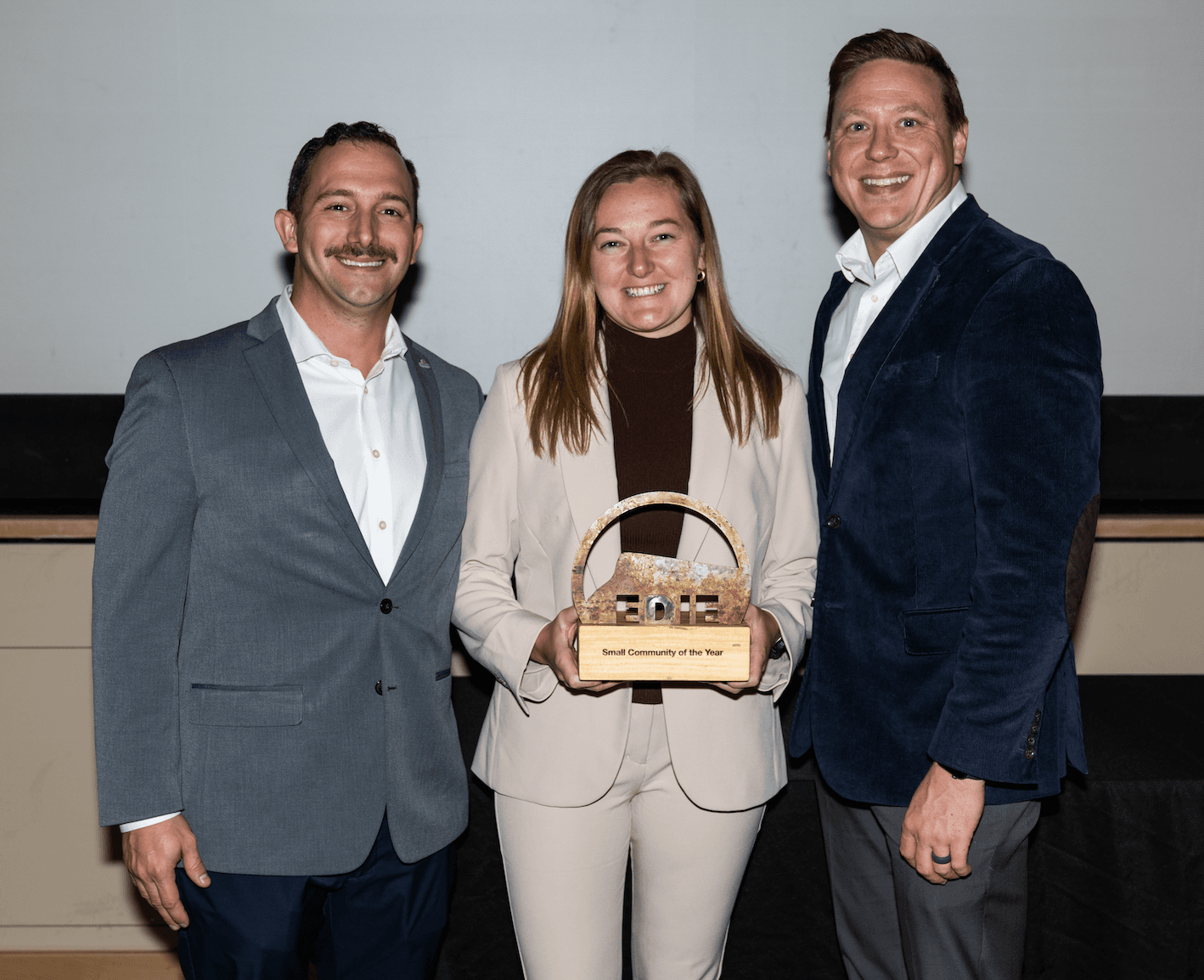 Three individuals in formal attire posing together, with the woman in the center holding an award plaque that reads "Small Community of the Year."
