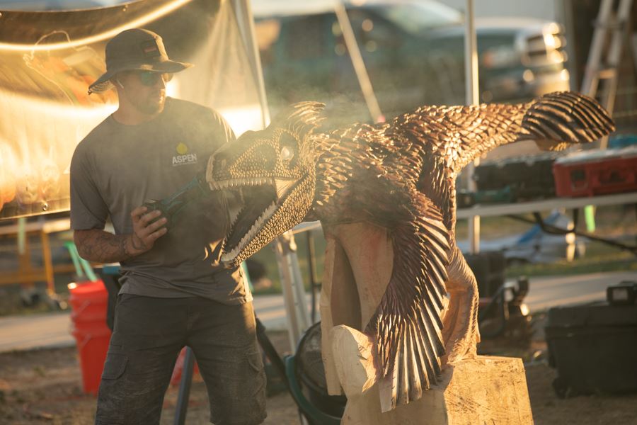 Artist carving a wooden dinosaur sculpture at an outdoor event.